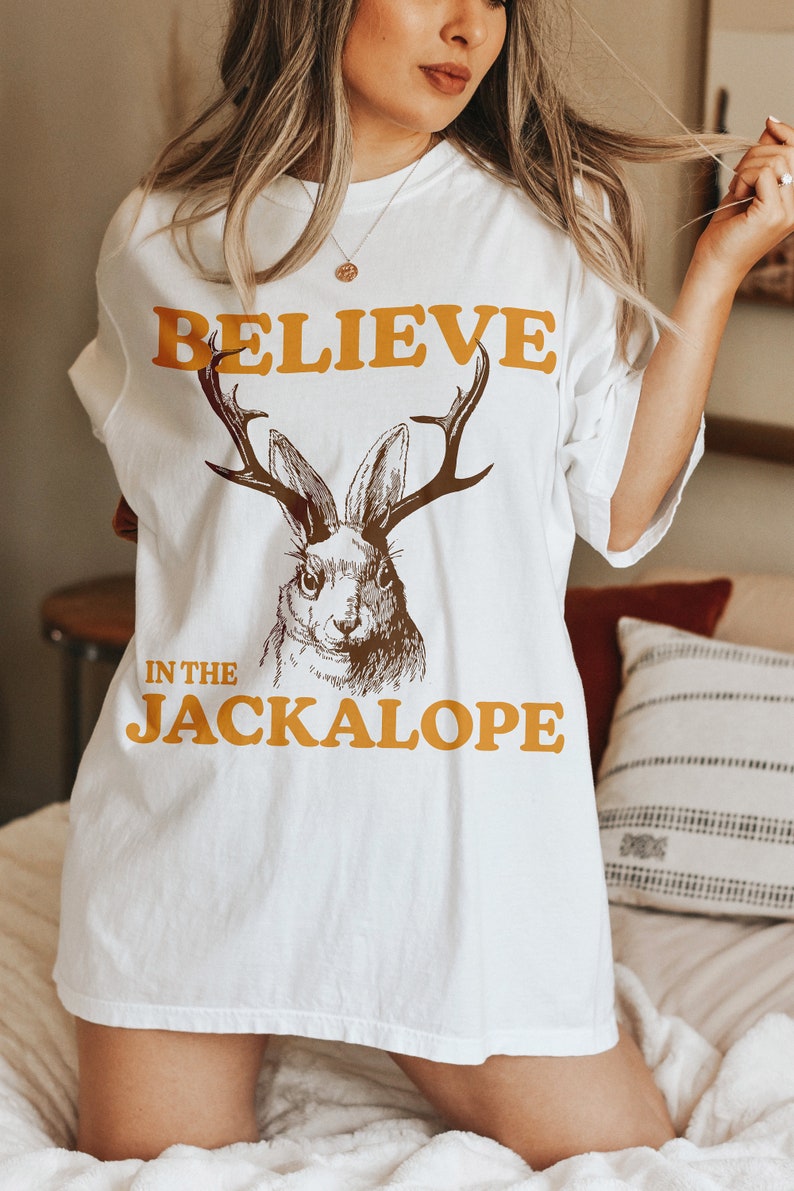 Pu&ograve; includere: Maglietta bianca con un'illustrazione marrone e bianca di un jackalope, un coniglio con le corna. Il testo "BELIEVE IN THE JACKALOPE" &egrave; stampato in arancione sulla maglietta.