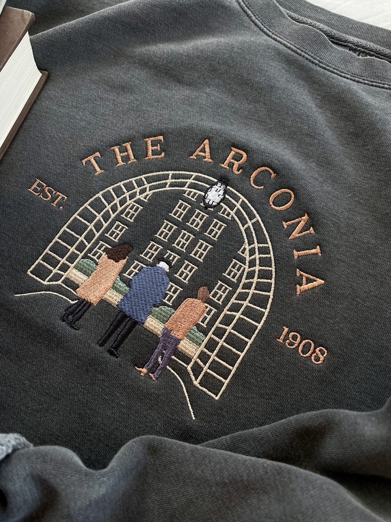 May include: A dark gray sweatshirt with an embroidered design featuring a building with windows, a clock tower, and four people standing in front of it. The text "THE ARCONIA EST. 1908" is embroidered above the building.