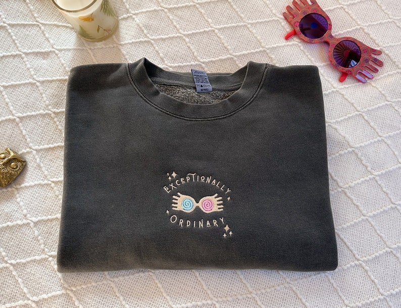 May include: A dark gray sweatshirt with a gold embroidered design featuring a pair of glasses with swirling pink and blue lenses and the text "Exceptionally Ordinary".