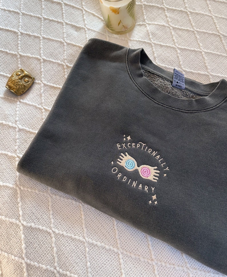 May include: A dark gray sweatshirt with a gold embroidered design. The design features a pair of glasses with swirling lenses and the text "Exceptionally Ordinary".