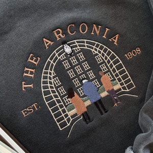 May include: A dark gray sweatshirt with an embroidered design featuring a building with windows, a clock, and three people standing in front of it. The text "THE ARCONIA EST. 1908" is embroidered around the design.