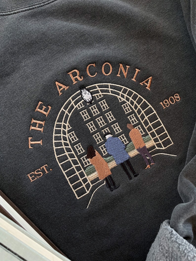 May include: A dark gray sweatshirt with an embroidered design featuring a building with windows, a clock, and three people standing in front of it. The text "THE ARCONIA EST. 1908" is embroidered around the design.