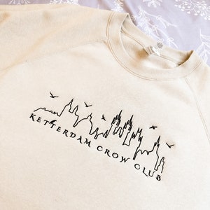 May include: A beige sweatshirt with a black embroidered design of a city skyline with the text "KETTERDAM CROW CLUB".