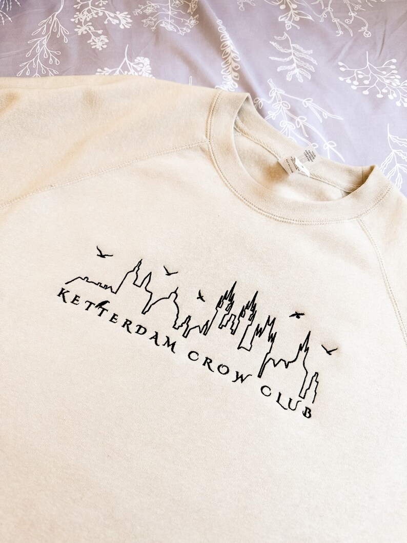 May include: A beige sweatshirt with a black embroidered design of a city skyline with the text "KETTERDAM CROW CLUB".