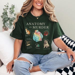 May include: A dark green t-shirt with the text "Anatomy of a Murder" printed on the front. The shirt features illustrations of a cat, a tie-dye hoodie, a red ring, a dog, and a green gem.