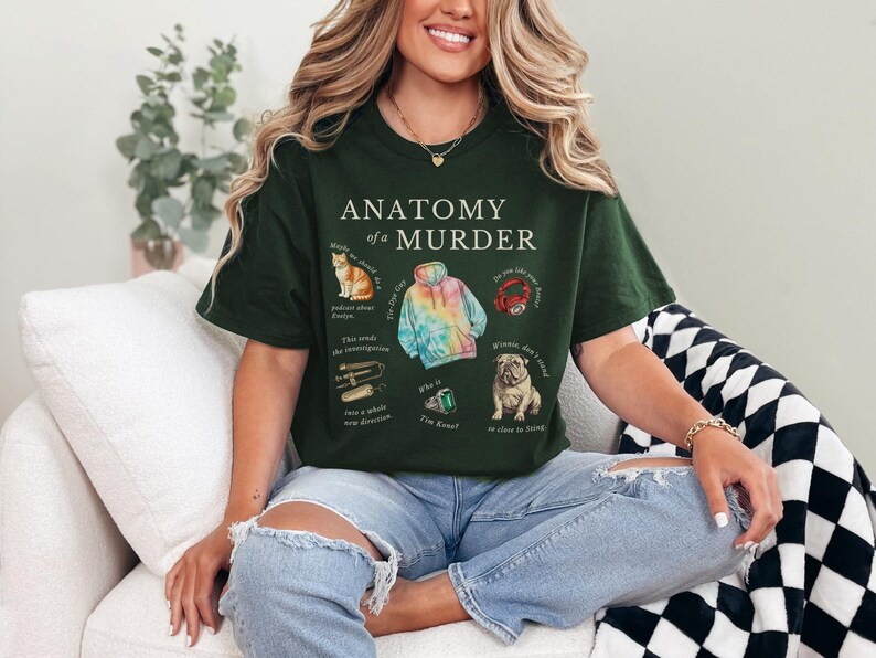 May include: A dark green t-shirt with the text "Anatomy of a Murder" printed on the front. The shirt features illustrations of a cat, a tie-dye hoodie, a red ring, a dog, and a green gem.