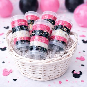 May include: A white wicker basket holds five push-pop containers. Each container is decorated with alternating bands of pink, black, and white glitter. The background includes pink and black balloons, confetti, and Minnie Mouse cutouts.