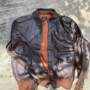 Könnte beinhalten: Braune Leder-Bomberjacke mit braunem Kragen, Bündchen und Bund. Die Jacke hat zwei Vordertaschen und einen Aufnäher am oberen linken Ärmel. Das Innenfutter ist hellbraun. Größe 38.