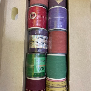 May include: A collection of colorful spools of thread, neatly stacked inside a cardboard box. The spools vary in color, including red, purple, green, and yellow. Some spools have labels with text. The thread appears to be tightly wound.