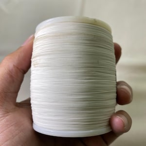 May include: A spool of white thread, tightly wound around a plastic core. The thread appears to be a fine, smooth material, suitable for sewing or crafting projects. The spool is held in a hand, showing the scale of the thread.