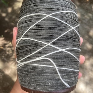 Könnte beinhalten: Eine große Spule dunkelgrauen Fadens mit weißem Faden, der sich über die Außenseite kreuzt. Die Spule hat einen dunkelbraunen Holzfuß und -deckel. Der Faden ist eng gewickelt und erzeugt eine strukturierte Oberfläche.