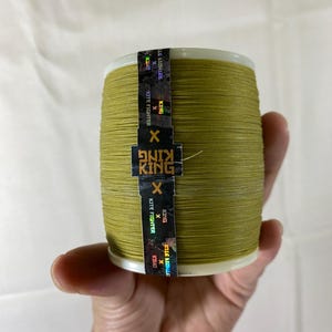 Puede incluir: Una mano sostiene una bobina de hilo de cometa verde oliva. La bobina tiene una base blanca y una etiqueta negra y dorada con el texto "KING" y "KITE FIGHTER".