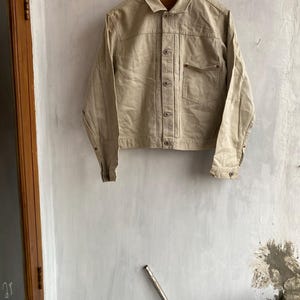 Op de afbeelding: Een beige spijkerjasje met een knoopsluiting, een kraag en een enkele borstzak. De jas hangt aan een houten hanger tegen een getextureerde witte muur. De jas heeft manchetten met knopen en een klassiek ontwerp.