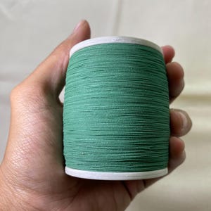 May include: A spool of green thread held in a hand. The thread is tightly wound around a white core. The thread is a solid, medium green color, suitable for sewing and crafting projects.