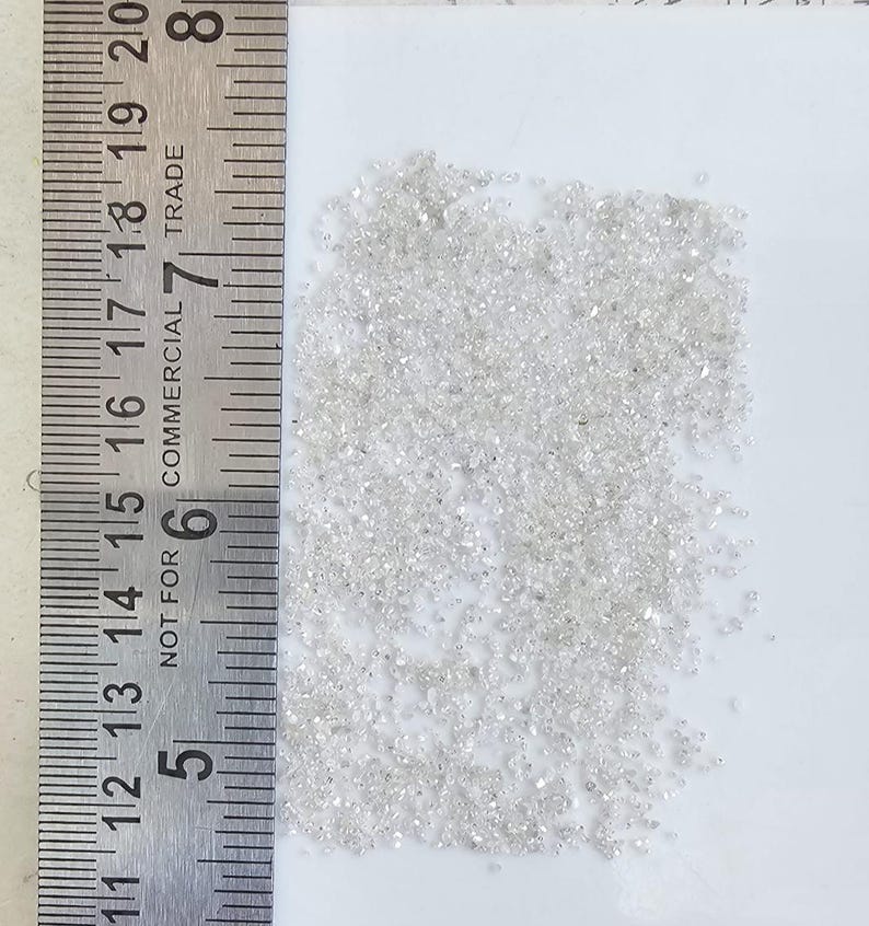 Puede incluir: Un mont&oacute;n de peque&ntilde;os diamantes transparentes junto a una regla met&aacute;lica. La regla muestra medidas en cent&iacute;metros, con los n&uacute;meros 28 a 50. Los diamantes est&aacute;n dispersos sobre una superficie blanca.