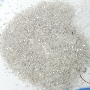 Puede incluir: Primer plano de un mont&oacute;n de material gris claro y brillante, similar a arena. Las peque&ntilde;as part&iacute;culas irregulares reflejan la luz, creando un efecto brillante. El material est&aacute; disperso sobre una superficie blanca, con algunas motas m&aacute;s oscuras.