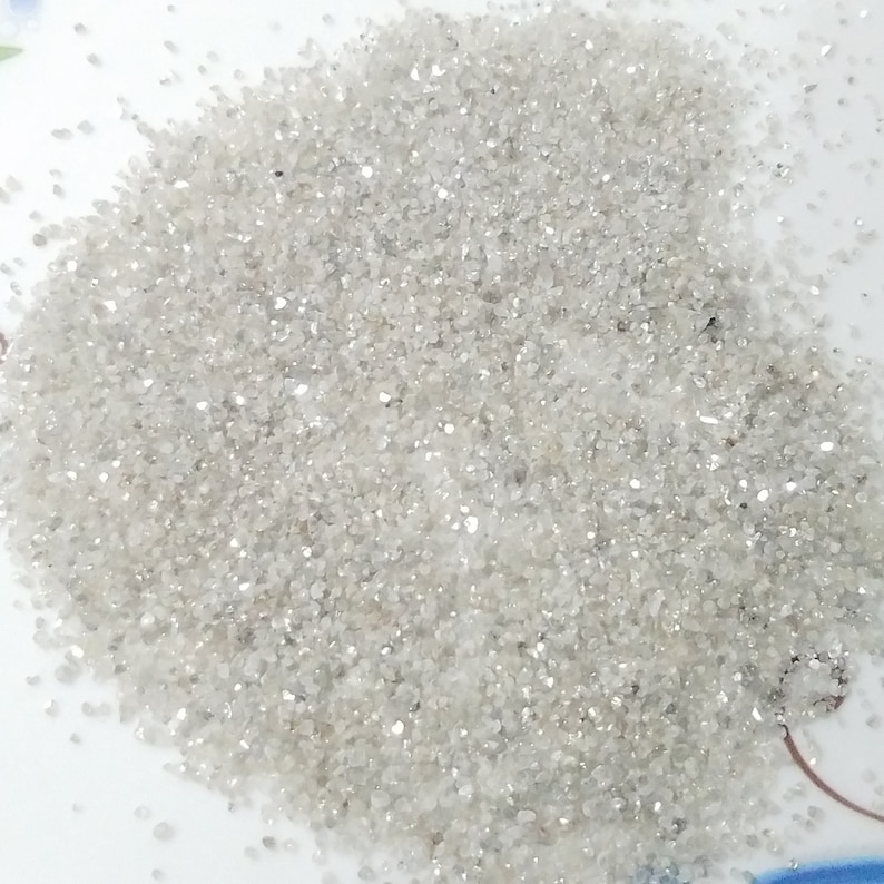 Puede incluir: Primer plano de un mont&oacute;n de material gris claro y brillante, similar a arena. Las peque&ntilde;as part&iacute;culas irregulares reflejan la luz, creando un efecto brillante. El material est&aacute; disperso sobre una superficie blanca, con algunas motas m&aacute;s oscuras.