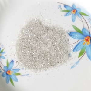 Puede incluir: Un mont&oacute;n de purpurina fina y brillante de color plateado sobre un plato blanco. El plato est&aacute; decorado con motivos florales azules. La purpurina parece estar distribuida uniformemente, captando la luz y creando un efecto brillante. Ideal para manualidades.