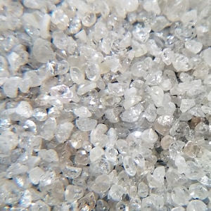 Puede incluir: Primer plano de un mont&oacute;n de peque&ntilde;as piedras preciosas claras e irregulares. Las piedras preciosas est&aacute;n dispersas, con la luz reflej&aacute;ndose en sus facetas, creando un efecto brillante. El color general es un blanco transl&uacute;cido.