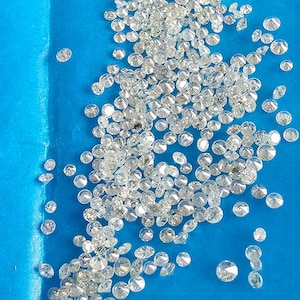 May include: A collection of small, clear diamonds scattered on a bright blue surface. The diamonds are round-cut and reflect light, creating a sparkling effect. The image is a close-up, showcasing the clarity and brilliance of the gemstones.