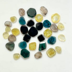 Può includere: Una collezione di gemme non tagliate in vari colori, tra cui giallo, verde acqua, verde scuro e nero. Le gemme hanno una forma irregolare e sono sparse su una superficie bianca. Le gemme hanno una superficie lucida e riflettente.