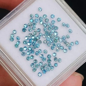 Op de afbeelding: Een verzameling kleine, ronde, lichtblauwe diamanten wordt gepresenteerd in een doorzichtige, vierkante plastic container. De diamanten zijn geslepen om te schitteren en licht te reflecteren. De container staat op een wit oppervlak.