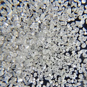 Puede incluir: Primer plano de una colecci&oacute;n de peque&ntilde;as gemas claras y facetadas. Las gemas est&aacute;n dispersas sobre una superficie oscura, reflejando la luz y creando un efecto brillante. Las formas var&iacute;an, lo que sugiere que son sin cortar o de corte basto.