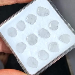 May include: A collection of twelve small, translucent, white, irregularly shaped gemstones arranged in a grid pattern inside a square, clear plastic display case with a white foam insert. Each stone features a faceted surface and light gray inclusions.