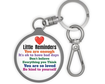 May include: A round keychain with a gray border and a silver clasp. The keychain features the text "Little Reminders" and inspirational phrases in various colors, including "You are enough" and "Be kind to yourself."