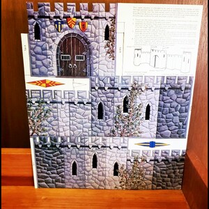 May include: A cardboard castle diorama with a stone-like facade, featuring a wooden door, arched windows, and decorative shields. The castle has a detailed design with printed instructions on the side. The diorama is set on a wooden surface.