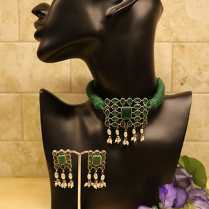 May include: A green choker necklace with a silver pendant featuring green stones and white pearls. Matching earrings with green stones and white pearls are displayed on a stand.
