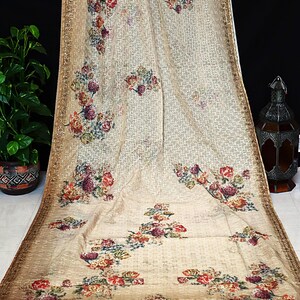 May include: A beige fabric wrap featuring a repeating geometric grid pattern and scattered floral clusters in shades of red, orange, purple, and green. The edges are finished with a decorative gold border and small tassels along the bottom hem.