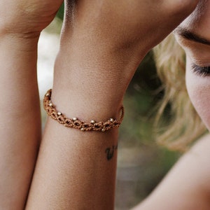 May include: A close-up of a person's arm wearing a brown braided bracelet with gold beads. The bracelet is on the left wrist and the person's right hand is resting on their forehead.