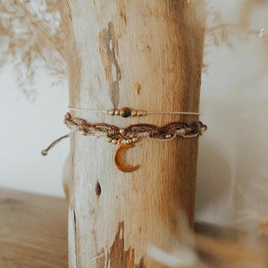 Könnte beinhalten: Zwei geflochtene Armbänder mit einem goldenen Halbmond-Anhänger. Die Armbänder sind aus braunem und weißem Garn gefertigt und haben kleine goldene Perlen.
