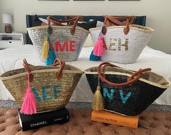 May include: Four straw tote bags with leather handles and colorful tassels. Each bag has a different name embroidered on the front: "AME", "AEH", "REF", and "NV". The bags are sitting on a brown leather bench with books on top.