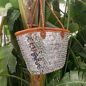Puede incluir: Un bolso tote de lentejuelas plateadas con asas y ribetes de cuero marrón. El bolso está colgado de una rama de árbol.