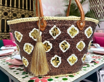 Peut inclure: Un sac cabas brun et or avec un pompon. Le sac est fait de paille tressée et a une finition scintillante. Le sac a une poignée en cuir brun et un pompon doré.