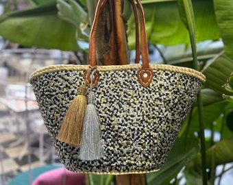 Puede incluir: Un bolso tote de paja negro y blanco con lentejuelas doradas y un borla dorado y plateado. El bolso tiene asas de cuero marrón.
