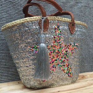 May include: A silver sequined tote bag with a star design made of colorful sequins. The bag has brown leather handles and a silver tassel. The bag is made of woven straw.