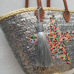 May include: A silver sequined tote bag with a star design made of colorful sequins. The bag has a woven straw top and handles with brown leather accents. A silver tassel hangs from the handle.