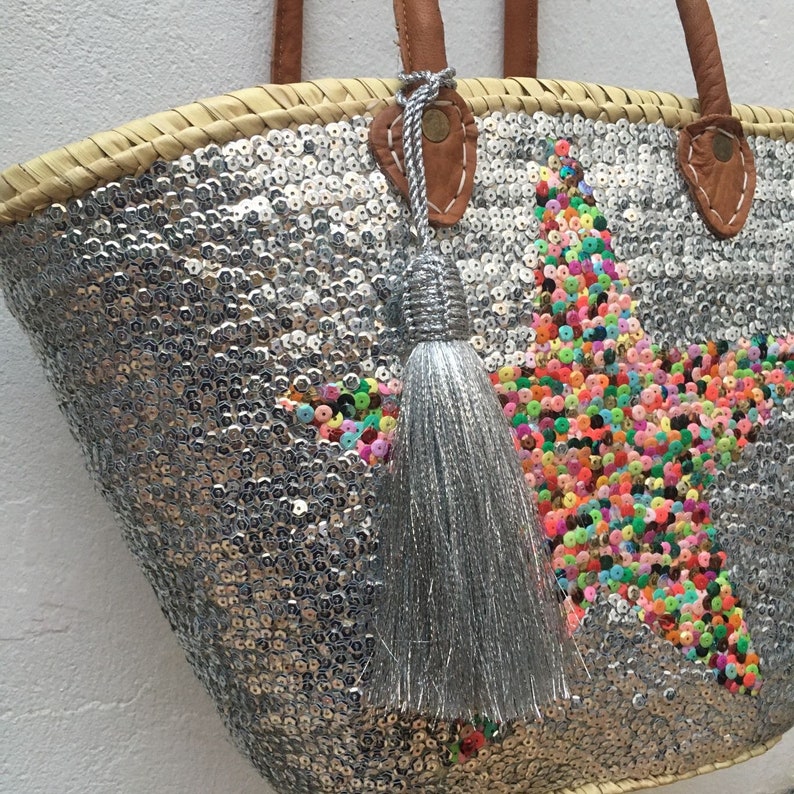 May include: A silver sequined tote bag with a star design made of colorful sequins. The bag has a woven straw top and handles with brown leather accents. A silver tassel hangs from the handle.