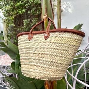 May include: A woven straw market bag with brown leather handles and trim. The bag is hanging from a tree branch.