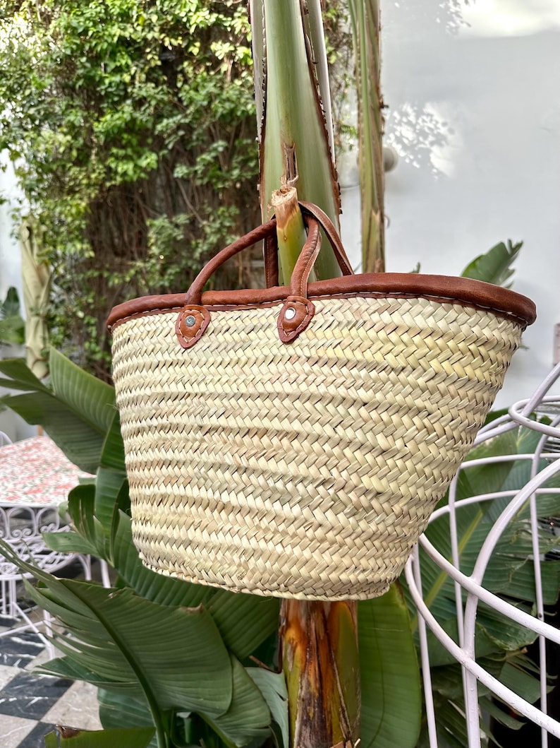 May include: A woven straw market bag with brown leather handles and trim. The bag is hanging from a tree branch.
