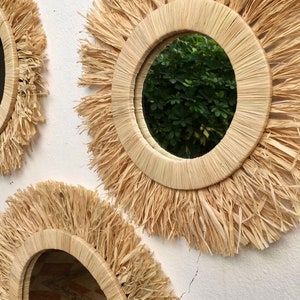 May include: Two round mirrors with a natural woven straw frame and a fringe border. The mirrors are hanging on a white wall.