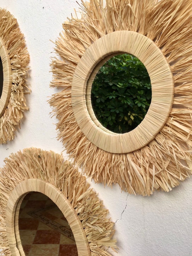 May include: Two round mirrors with a natural woven straw frame and a fringe border. The mirrors are hanging on a white wall.