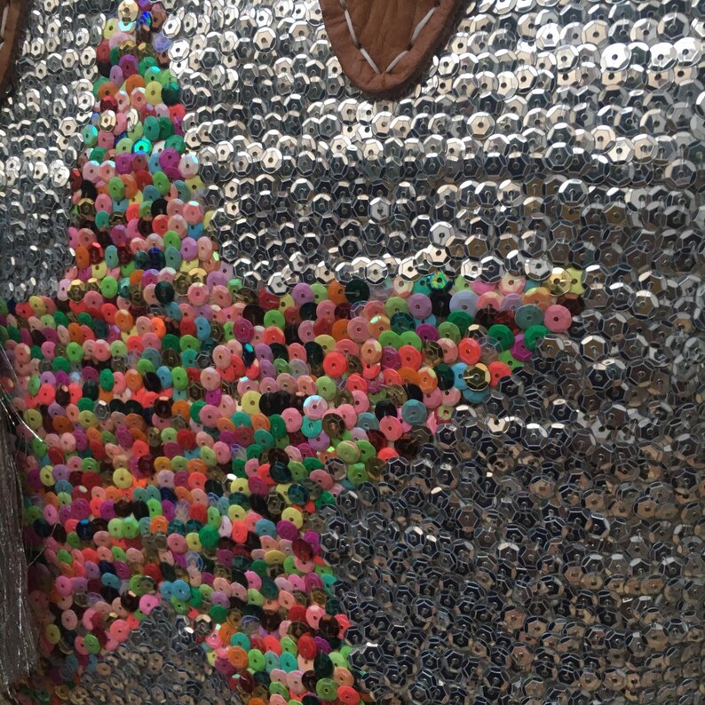 May include: Silver sequined handbag with a star-shaped design made of colorful sequins. The bag has a brown leather handle with white stitching. The sequins are in various colors, including pink, green, orange, and yellow.