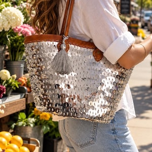 Peut inclure: Un sac cabas recouvert de sequins argentés réfléchissants avec des anses et des bordures en similicuir marron. Un pompon gris décoratif est suspendu à l'une des anses. Le sac présente une forme rectangulaire avec une large ouverture.