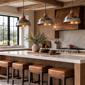 May include: Three dome-shaped pendant lights with a weathered copper and teal patina finish hang above a kitchen island. The island features a white marble countertop and four square stools with brown leather seats and dark wooden frames.