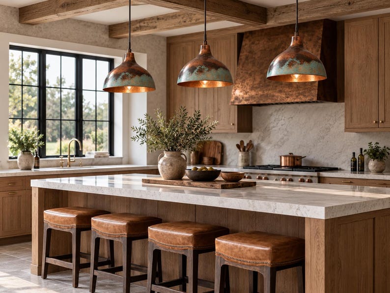 May include: Three dome-shaped pendant lights with a weathered copper and teal patina finish hang above a kitchen island. The island features a white marble countertop and four square stools with brown leather seats and dark wooden frames.