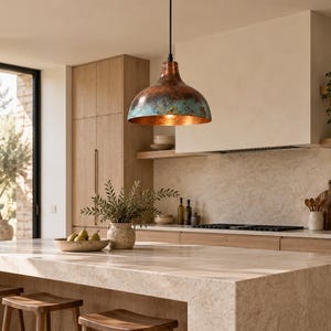 May include: A dome-shaped pendant light fixture featuring a metallic copper exterior with a weathered, oxidized teal patina finish. The interior of the shade is polished copper, suspended from a thin black cord centered above a kitchen island.